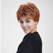 A smiling woman with vibrant blue eyes, centered against a plain grey background, poses and looks back over her shoulder. She wears a short, layered auburn pixie cut wig with copper highlights. Her black cardigan frames her upper body, and long, gold dangle earrings with pearls are visible. The lighting is even and soft.
