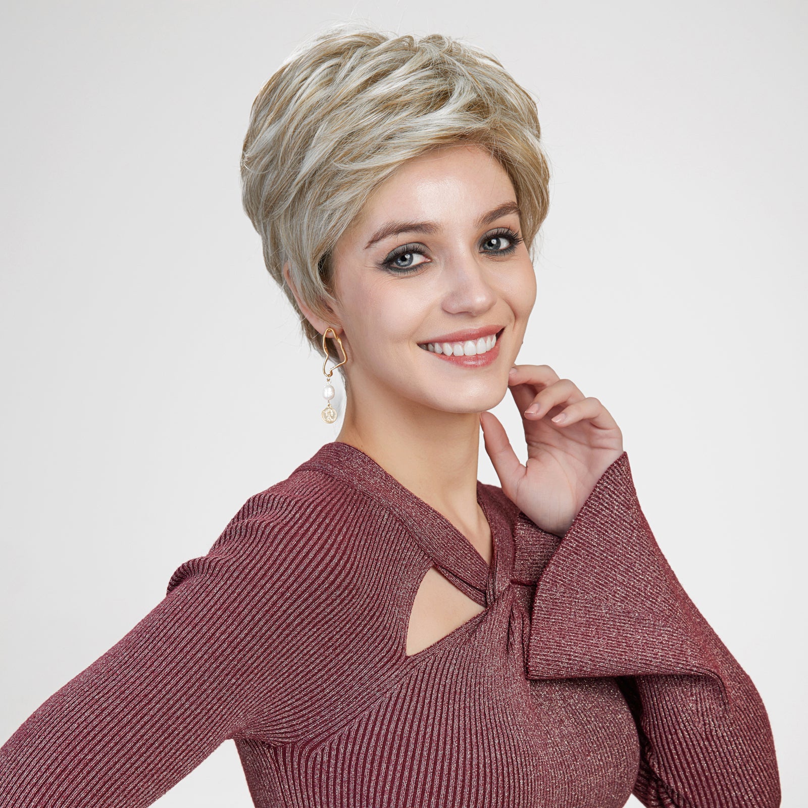 A studio photograph of a smiling woman with vibrant blue eyes, centered against a plain grey background. She wears a textured, shimmering burgundy long-sleeved top with a keyhole cutout at the chest. Her short, voluminous, layered pixie cut wig is a mix of light blonde and darker ash brown tones. A gold earring with a hanging pearl and coin charm is visible on her left ear. Her right hand is gently touching her neck. The lighting is bright and even.