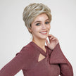 A studio photograph of a smiling woman with vibrant blue eyes, centered against a plain grey background. She wears a textured, shimmering burgundy long-sleeved top with a keyhole cutout at the chest. Her short, voluminous, layered pixie cut wig is a mix of light blonde and darker ash brown tones. A gold earring with a hanging pearl and coin charm is visible on her left ear. Her right hand is gently touching her neck. The lighting is bright and even.