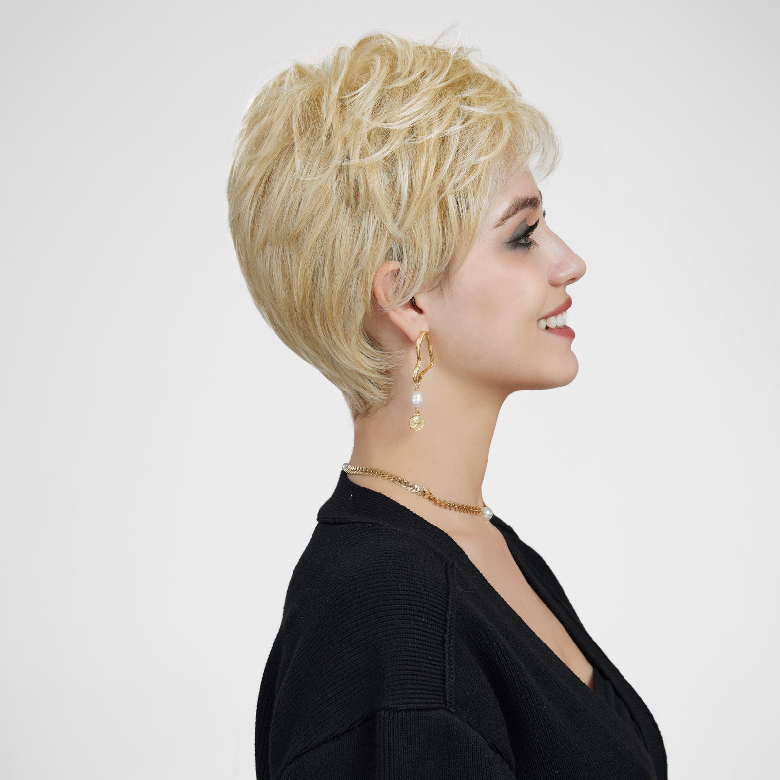 A side-profile studio portrait of a smiling woman against a plain grey background, modeling a short, layered blonde pixie cut wig. She wears a black cardigan, gold drop earrings, and a gold necklace.
