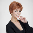 A studio portrait photograph of a smiling woman with blue eyes against a plain grey background. She models a short, layered auburn and copper pixie cut wig with highlights. She wears a black cardigan, a delicate pearl and gold choker necklace, and matching gold drop earrings with pearls. Her right hand touches her cheek. The lighting is even and soft.