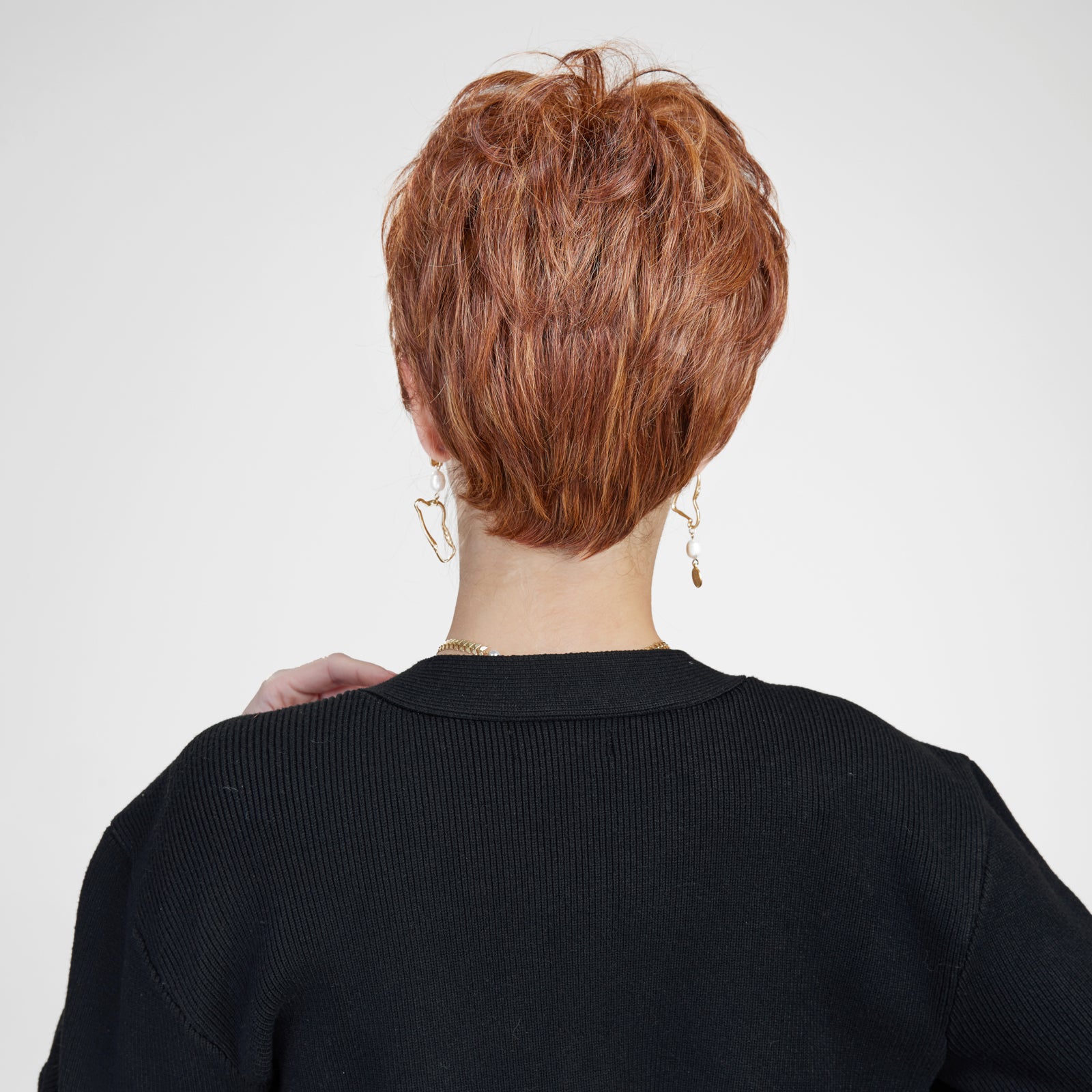 A back-view photograph of a woman modeling a short, layered auburn pixie cut wig with dimensional copper highlights. The wig has volume at the crown and tapers neatly at the nape, showcasing its textured layers. She wears a black ribbed cardigan and long, gold dangle earrings with small white pearls. Her hand is gently touching her neck. The background is a plain light grey. The lighting is soft and even.
