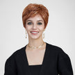 A studio portrait photograph of a smiling woman with blue eyes against a plain grey background, modeling a short, layered auburn and copper pixie cut wig with highlights. She wears a black cardigan, a delicate pearl and gold choker necklace, and matching gold drop earrings with pearls. The lighting is even and soft.