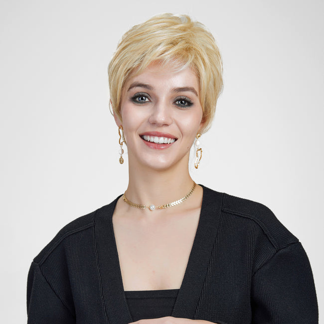 A smiling woman with blue eyes against a plain grey background, modeling a short, layered blonde pixie cut wig. She wears a black cardigan, gold earrings, and a delicate gold and pearl choker.