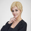 A smiling woman with blue eyes against a plain grey background, modeling a short, layered blonde pixie cut wig. She wears a black cardigan, gold earrings, and a gold and pearl choker necklace.