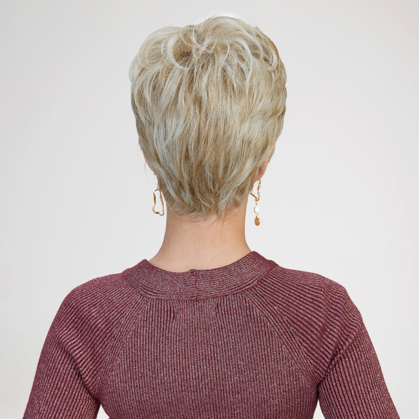 A back view photograph of a woman modeling a short, layered blonde pixie wig. The hair features dimensional blonde shades, blending highlights and lowlights for a natural look. The wig is styled with volume at the crown and tapers neatly at the nape of the neck, showing the distinct, textured layers. The woman is wearing a textured, shimmering burgundy long-sleeved top and small gold dangle earrings. The background is a plain, seamless light grey. The lighting is soft and even.