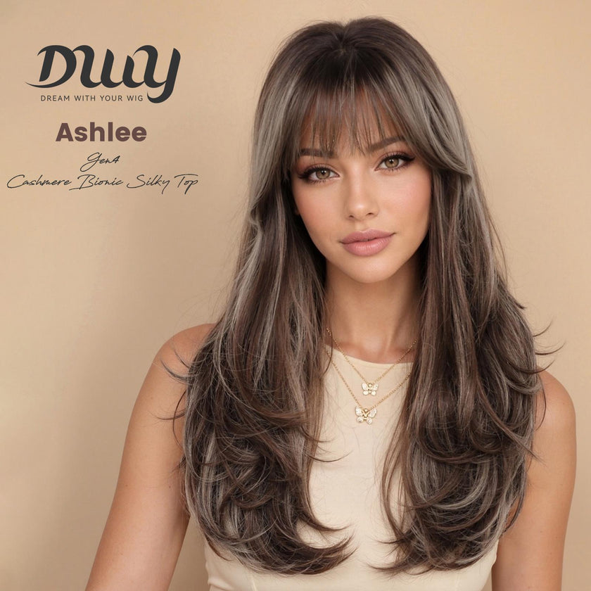 A woman wearing a beige top models the DWY Ashlee Gen4 synthetic wig, featuring long layered brown hair with blonde highlights, bangs, and a natural look.