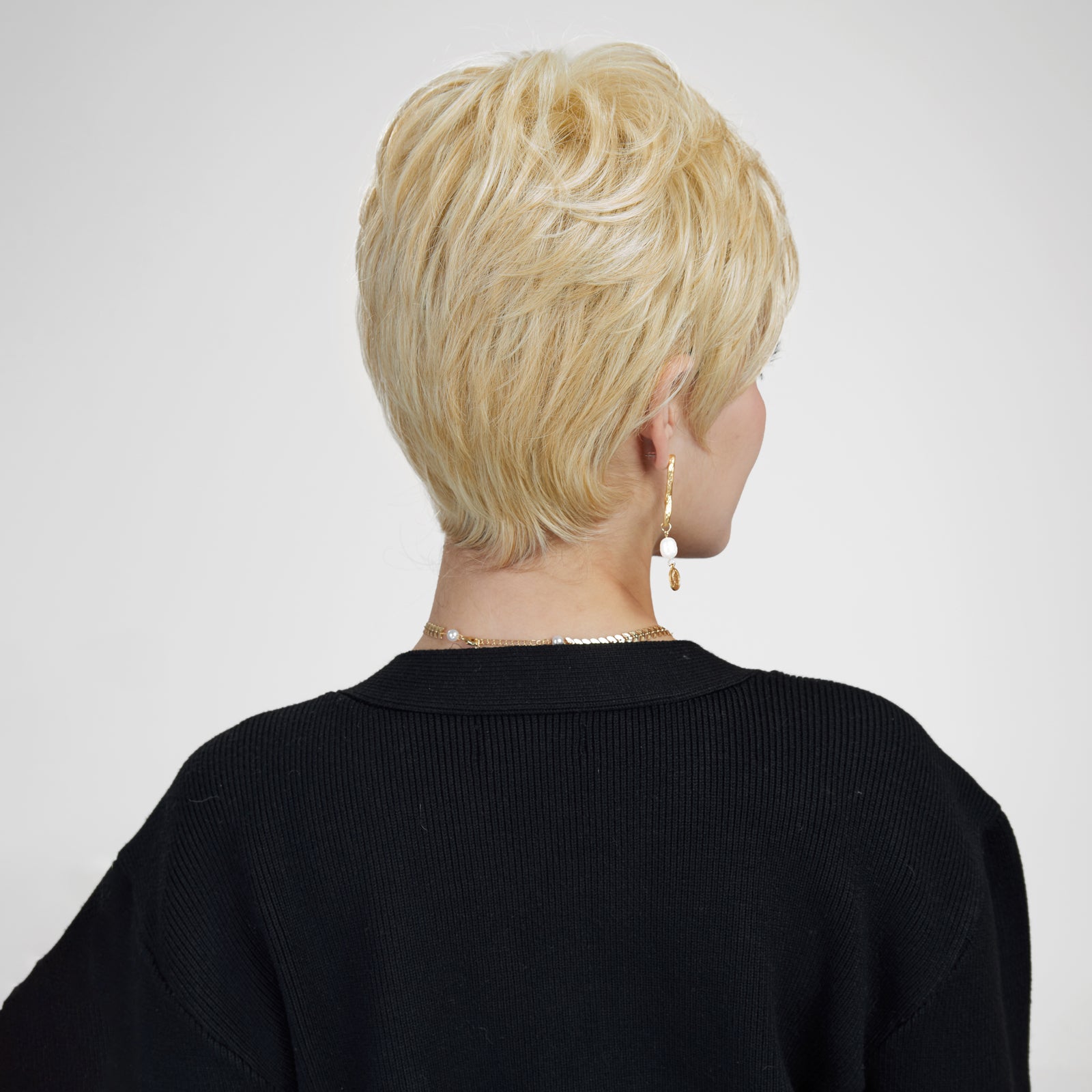 A back-view photograph of a woman against a plain grey background, modeling a short, layered blonde pixie cut wig. She wears a black cardigan, gold chain necklace, and white and gold dangle earrings. The wig features voluminous layers at the crown and a tapered nape. The lighting is soft and even.