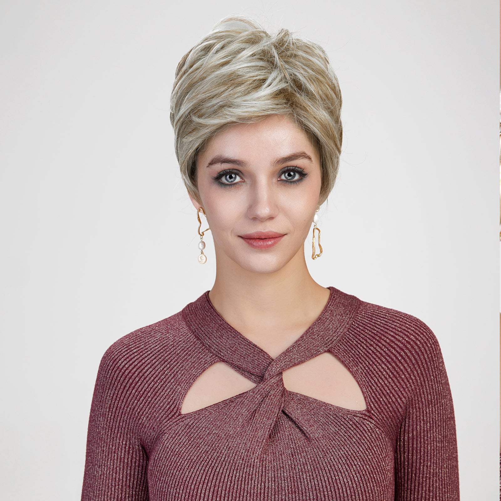 A studio portrait of a smiling woman against a grey background, modeling a short, layered blonde pixie wig.