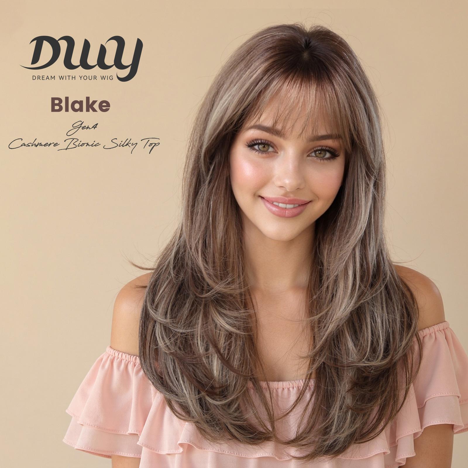 A woman with long, layered ash-brown and blonde highlighted hair with bangs, wearing a pink top, models a DWY Blake Gen4 Cashmere Bionic Silky Top synthetic wig against a beige background.