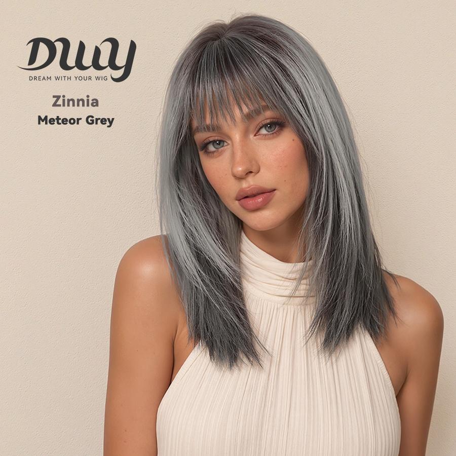 A candid studio photograph of a woman modeling a long, straight, layered wig with full bangs. The hair color is a multi-tonal, cool-toned Meteor Grey, darker towards the roots and transitioning to a silver-grey. She wears a cream-colored pleated halter top. In the top left corner, there is a dark grey logo with a stylized text that reads 