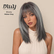 A candid studio photograph of a woman modeling a long, straight, layered wig with full bangs. The hair color is a multi-tonal, cool-toned Meteor Grey, darker towards the roots and transitioning to a silver-grey. She wears a cream-colored pleated halter top. In the top left corner, there is a dark grey logo with a stylized text that reads 
