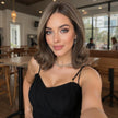 A close-up portrait of a beautiful woman with light brown hair and green eyes, smiling at the camera in a restaurant. She wears a stylish black DWY dress and delicate jewelry. The blurred background shows tables and blurred figures.