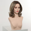 A realistic female mannequin head, set against a plain white studio background, displays a medium-length DWY wig. The layered hair is a blend of dark brown and ash brown, styled with a central part and flipped-out, voluminous curls at the ends. The mannequin wears subtle makeup and is positioned from a 3/4 view.