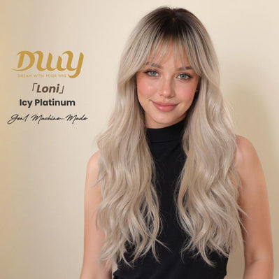 A medium shot photograph of a smiling woman modeling a long, wavy, icy platinum blonde wig with a dark root and curtain bangs. She is positioned slightly to the right against a plain beige studio background. In the upper left corner, there is gold text branding that reads 